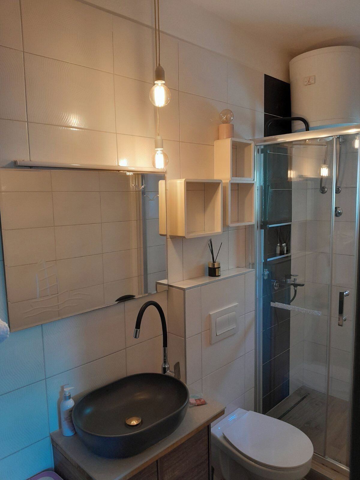 A modern bathroom with a large mirror, a black sink, a toilet, and a glass-enclosed shower.