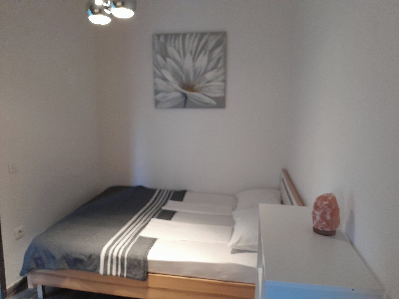 A small, neatly made bed with a striped blanket and white pillows, a small table with a salt lamp, and a wall-mounted abstract art piece.