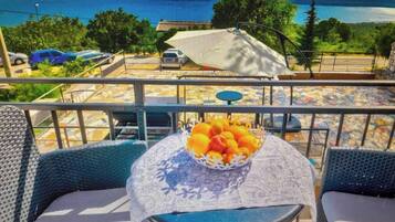 A balcony with a table set for two, a bowl of fruit, and a view of a parking area and trees.