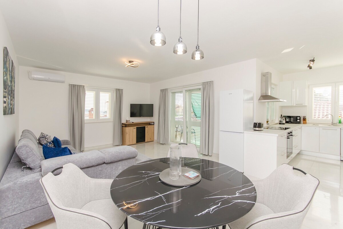 A modern apartment with a dining area featuring a marble table, a grey sofa with cushions, and a kitchen with white cabinets and a built-in oven.