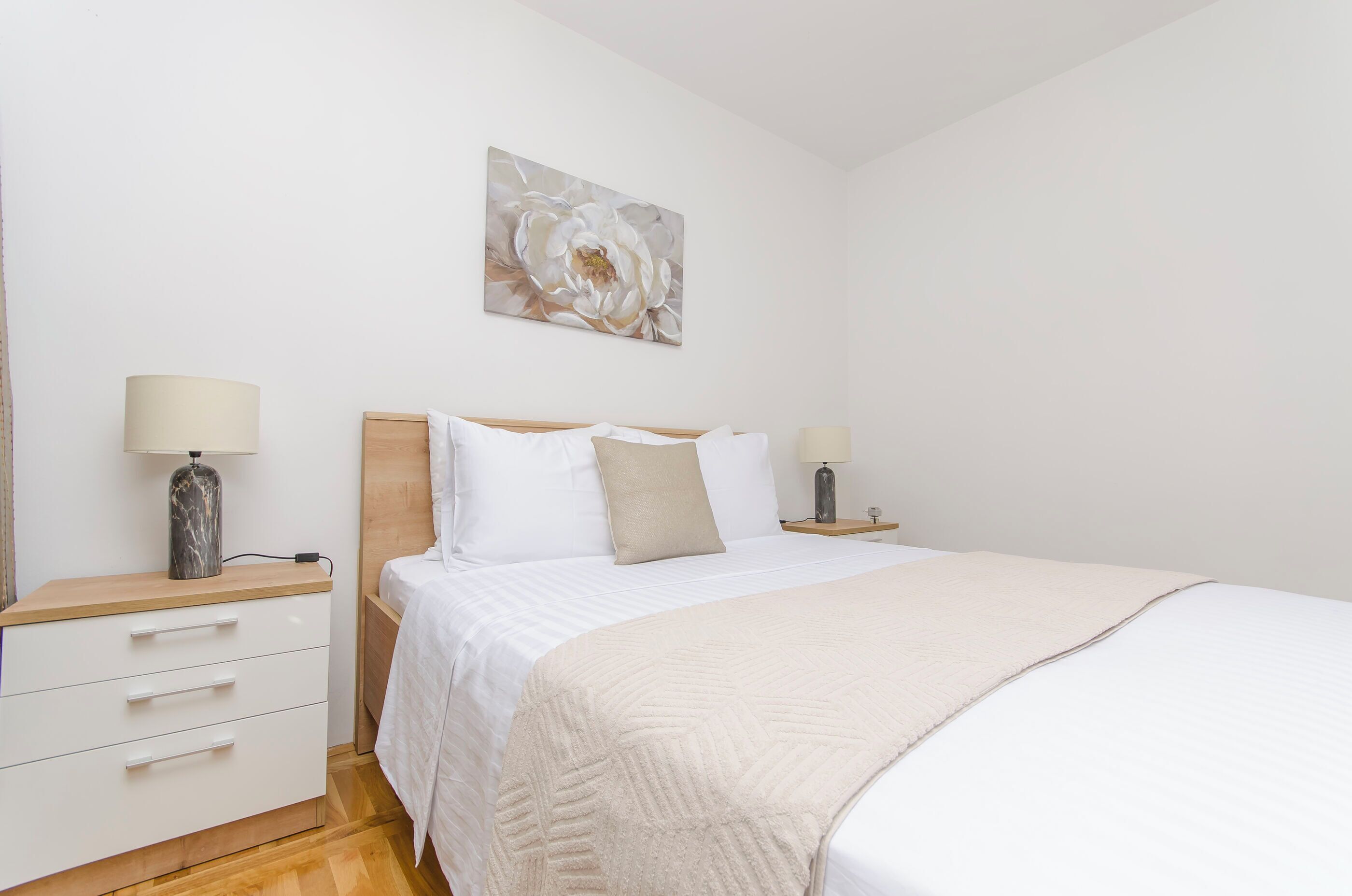 A bedroom with a bed, two bedside tables, a nightstand, a lamp, a drawer unit, and a wall-mounted painting.