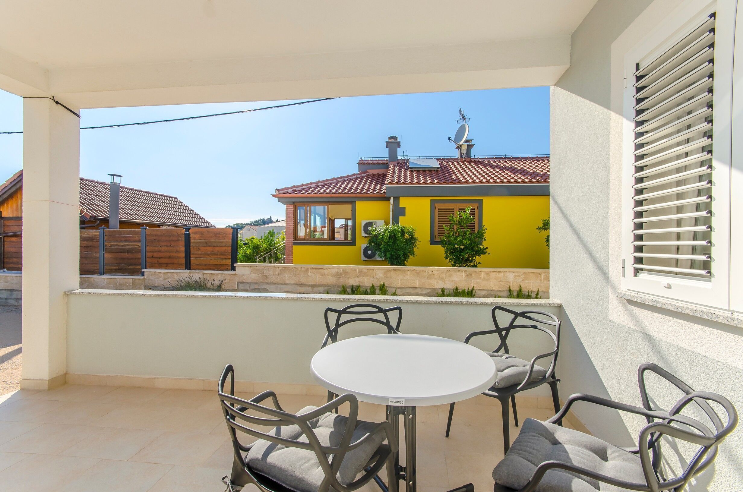 Ein Balkon mit einem weißen Tisch und vier Stühlen, ein gelbes Gebäude im Hintergrund und ein Fenster mit Jalousien.