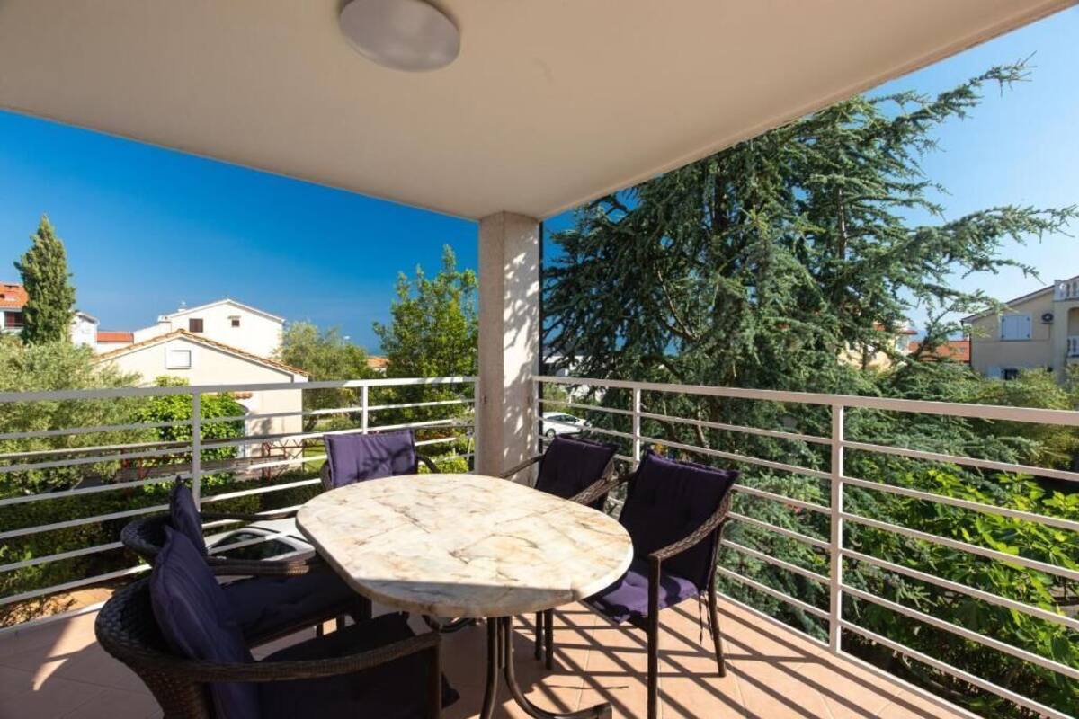 Un balcón con una mesa redonda y sillas, rodeado por una barandilla blanca, con vistas a una zona residencial con árboles y casas.