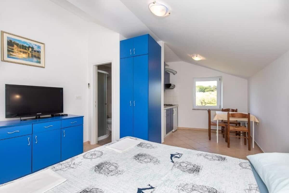 Un dormitorio con cama, televisor, armario azul, zona de comedor con mesa y sillas, y ventana con vistas.