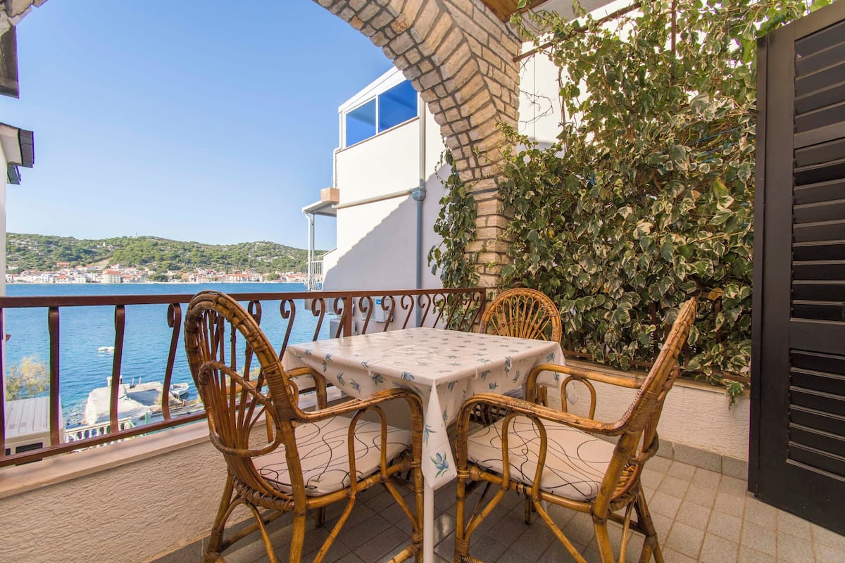 A balcony with wicker chairs and a table, overlooking a body of water and a distant town.
