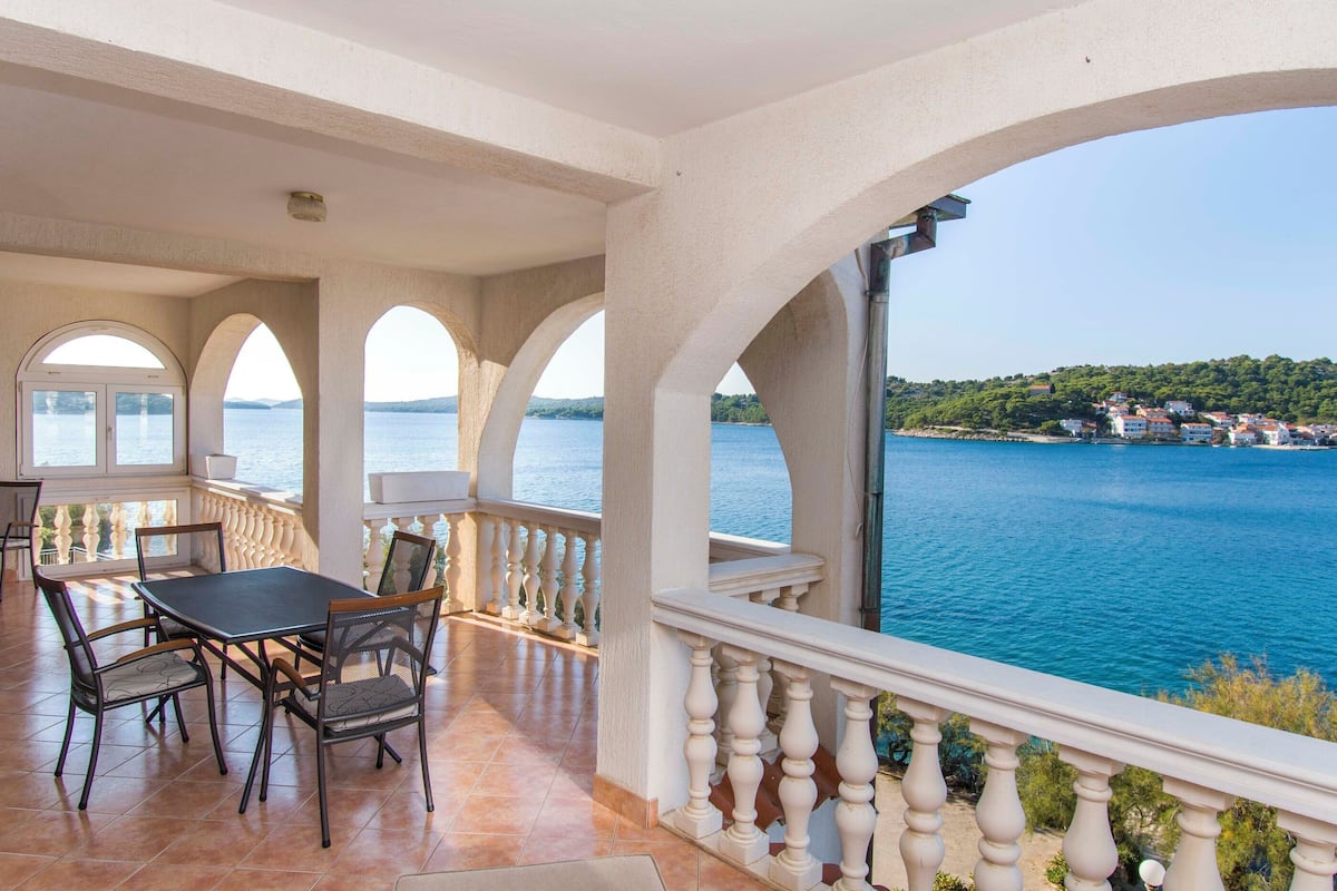 A covered patio with a table and chairs, overlooking a body of water and a distant coastline.