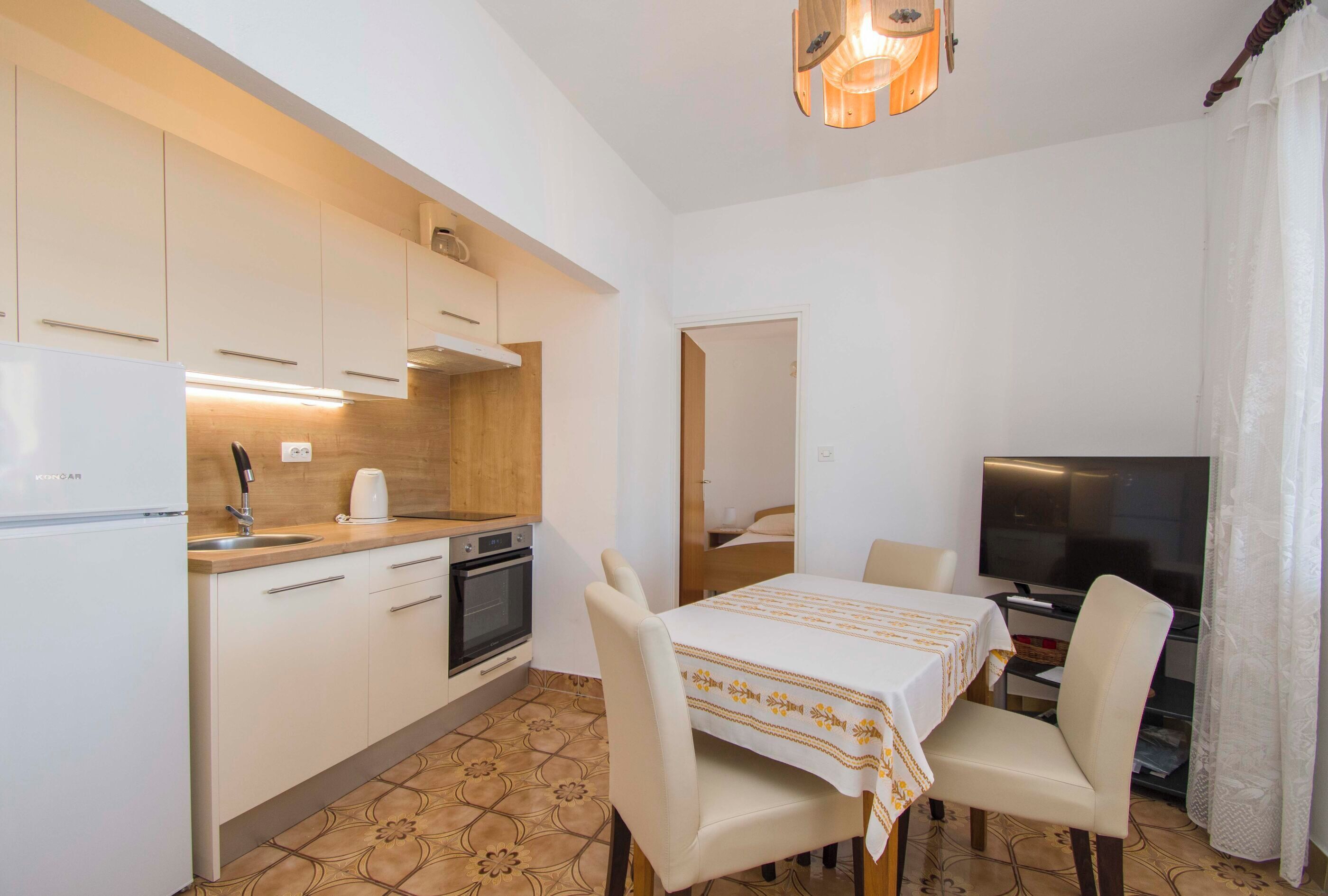 Cocina compacta con gabinetes blancos, horno empotrado y un lavamanos. Hay una mesa de comedor con sillas y un televisor en la esquina.