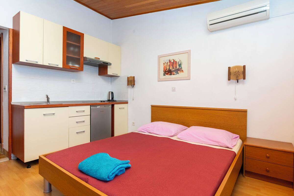 Un dormitorio con una cama de madera, una colcha roja y una toalla azul. Incluye un mesita de noche de madera y un cajón pequeño. Las paredes son blancas, el techo es de madera y hay un equipo de aire acondicionado instalado en la pared.