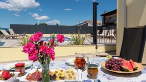 A table with food and drinks, a vase of flowers, and a view of a swimming pool and buildings. - Apartments Neda (Bilice)