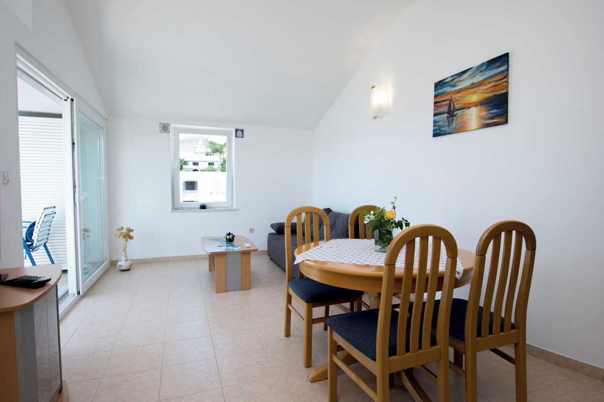 A dining area with a round table, four wooden chairs, a small table with a vase of flowers, and a painting on the wall.