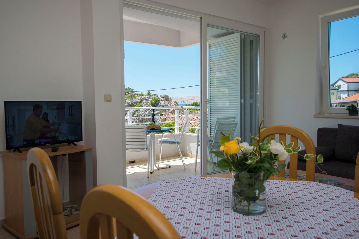 A living room with a television, a dining table with chairs, a vase with flowers, and a balcony with a view of the sea.