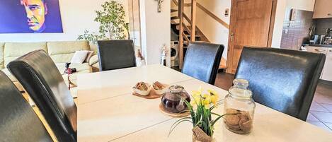 Ein Essbereich mit Tisch für zwei Personen, inklusive einer Vase mit Blumen, einer Glasdose und Brot. Eine Treppe führt in einen anderen Raum, und ein Fernseher ist an der Wand montiert.