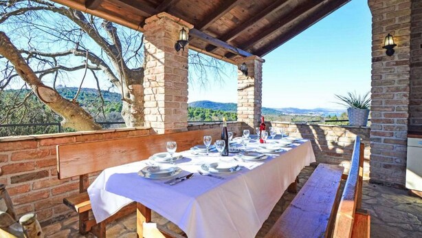 An outdoor dining area with a table set for six, stone pillars, wooden benches, and a view of the countryside. - Apartment Bilice (Bilice)
