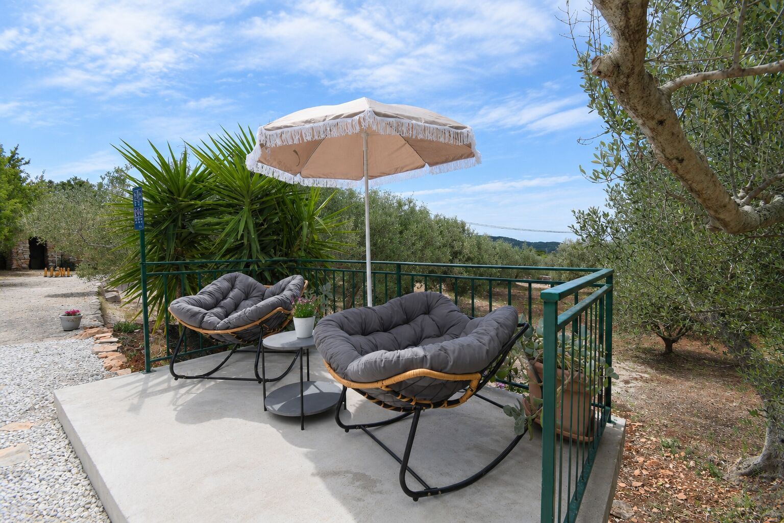 Ein Sitzbereich im Freien mit zwei gepolsterten Stühlen, einem Tisch und einem großen Sonnenschirm, sowie einem Metallgeländer.