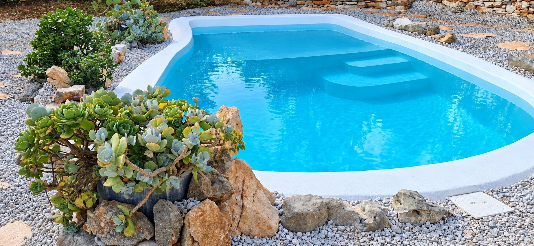 Une piscine rectangulaire bordée de blanc, entourée de rochers et de petites plantes.