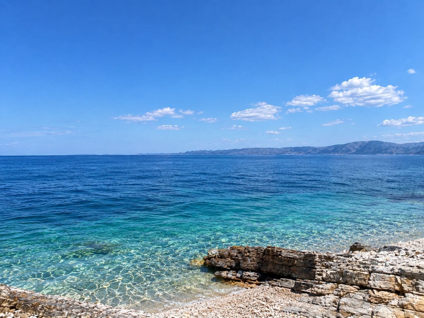 A coastal landscape with clear blue sea, rocky shoreline, and distant mountains.