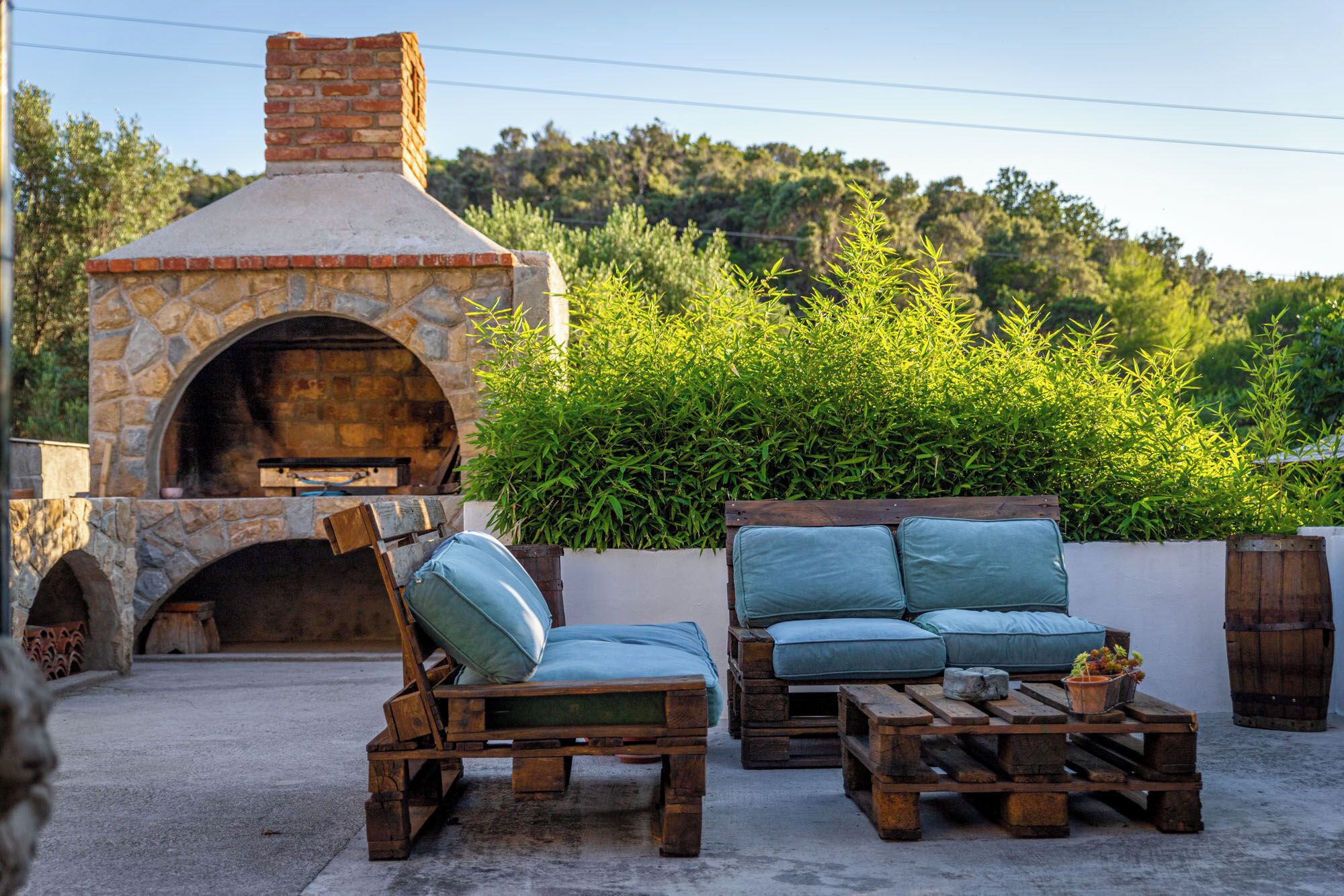 An outdoor seating area with stone and wooden furniture, a stone oven, and a view of a forested hillside.