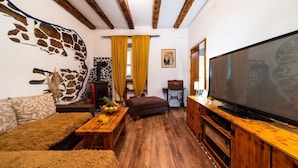 A cozy living room with a wooden floor, a sofa, a coffee table with fruit, a TV on a wooden stand, and a giraffe mural on the wall. - House Garden House (Rab)