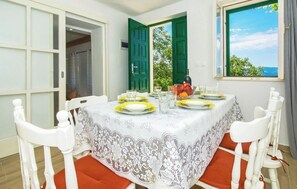 A dining area with a table set for four, white chairs, a lace tablecloth, and a view of greenery and the sea through a window. - House Summer Dream With Jacuzzi (Omis)