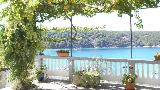 A balcony with potted plants, a white railing, and a view of a lake with boats. - Apartment Kampor (Rab)