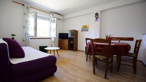 A living room with a purple sofa, a dining table with chairs, a small round table, a television, and a cabinet. - Apartment Kampor (Rab)