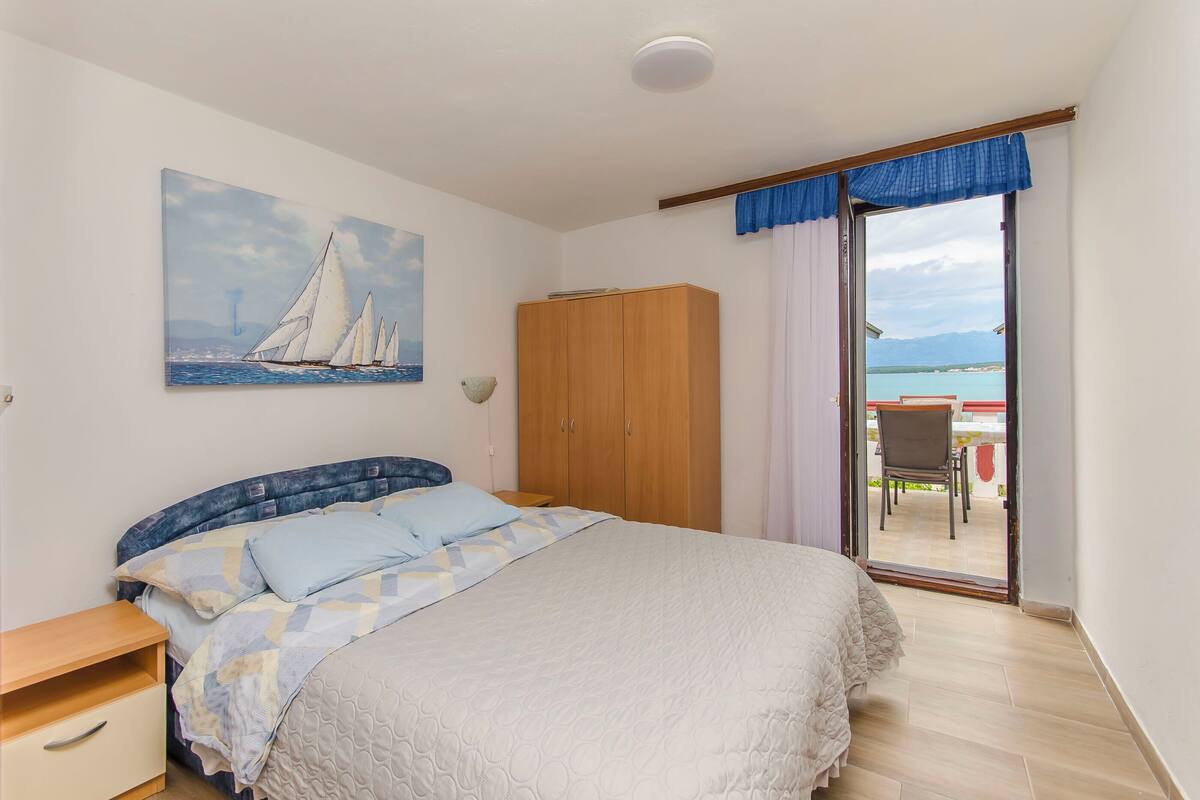Une chambre à coucher avec un lit, une armoire, un tableau représentant des voiliers et une vue sur la mer, visible à travers une porte coulissante.