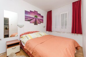 A bedroom with a bed, a dresser, a mirror, a window with red curtains, and a wall with a lavender field painting. - Apartment Kipson (Nin)