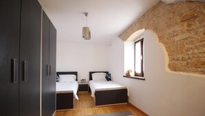 Una habitación con dos camas, piso de madera, alfombra con aro de hula y pared de piedra, además de una ventana con un osito de peluche.