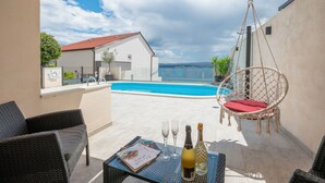A terrace with wicker furniture, a hanging chair, a table with a bottle and glasses, and a pool in the background. - House Mimica (Omis)