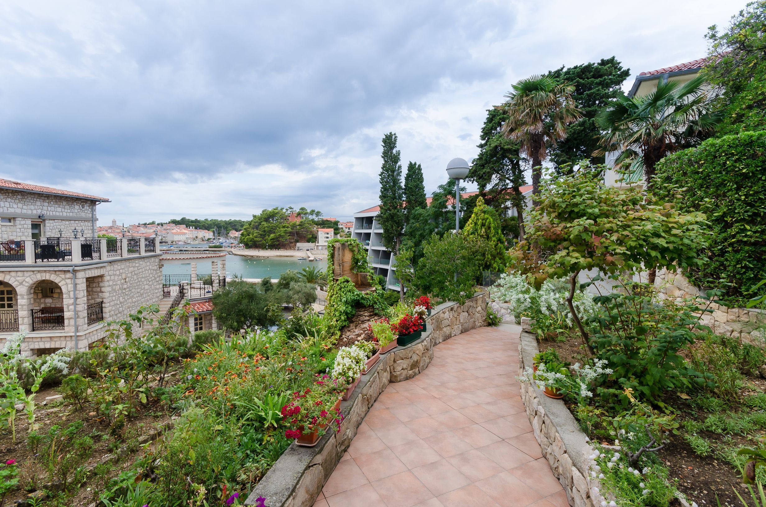 Un sentier de jardin mène à un bâtiment en pierre, d’où l’on peut admirer un plan d’eau et la verdure environnante.