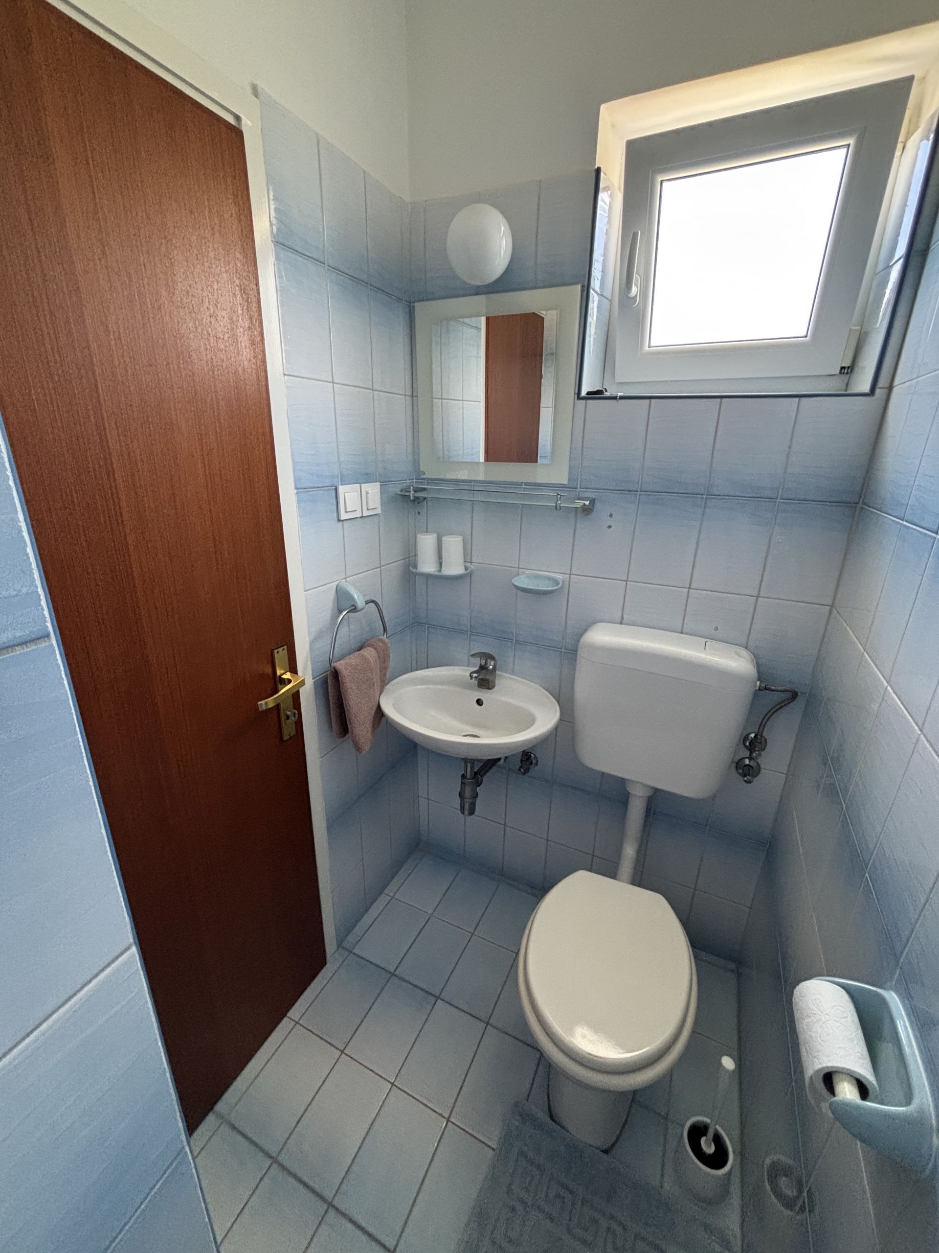 A compact bathroom with a toilet, sink, and a small window above the mirror.