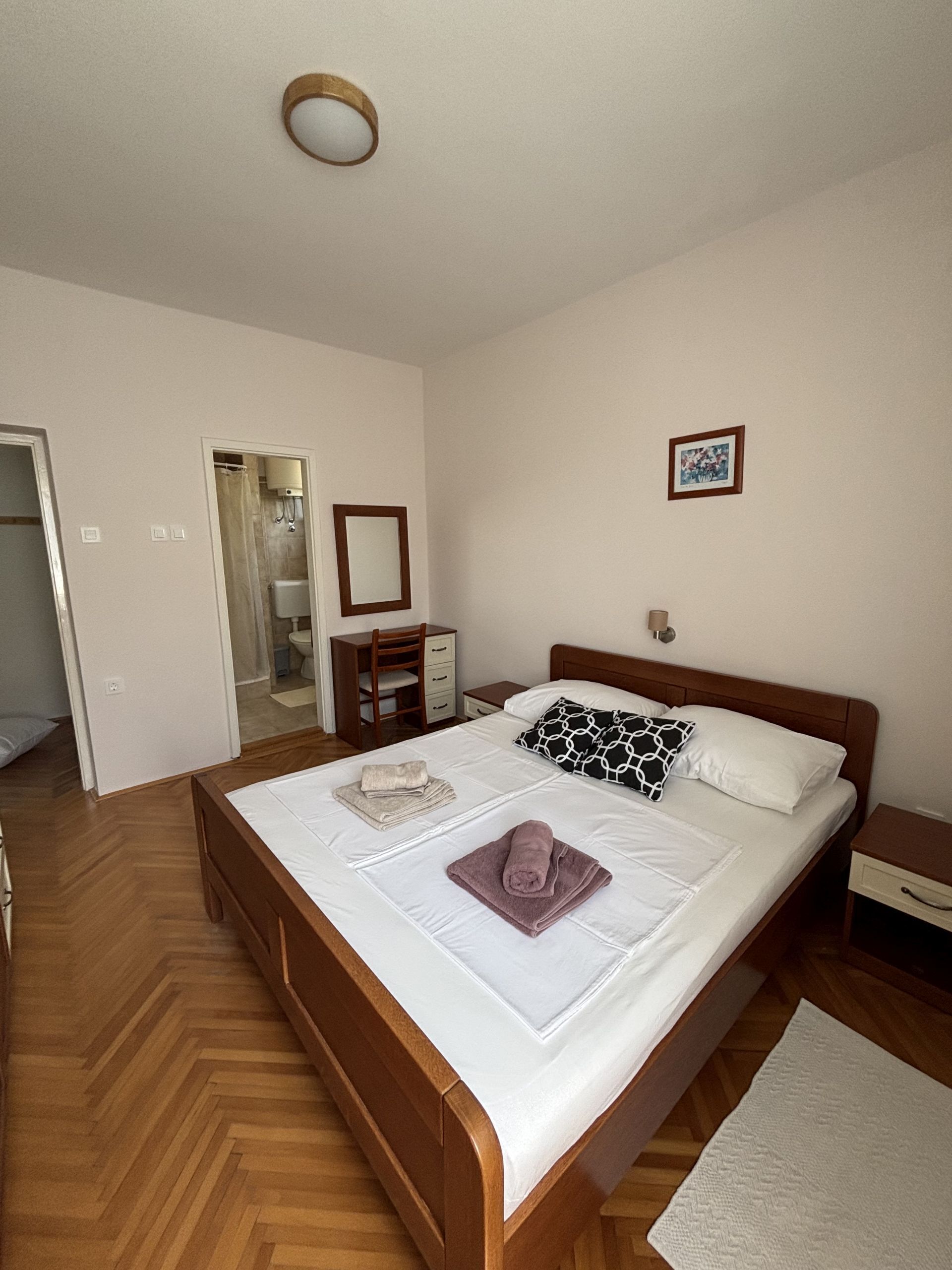 A neatly arranged bedroom with a wooden bed, a bedside table, a nightstand, a mirror, and a framed picture on the wall.