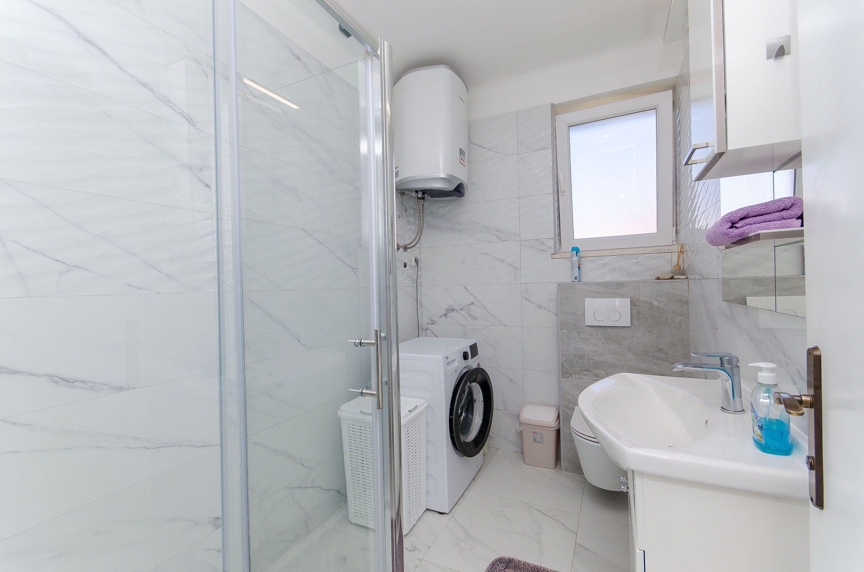 A bathroom with a glass shower enclosure, a washing machine, a sink, and a window.