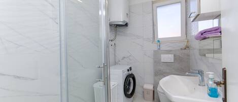 A bathroom with a glass shower enclosure, a washing machine, a sink, and a window.