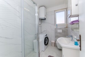 A bathroom with a glass shower enclosure, a washing machine, a sink, and a window. - Apartment Sun City (Split)