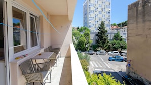 A balcony with a table and chairs, a view of a street with cars, and a tall building in the background. - Apartment Sun City (Split)