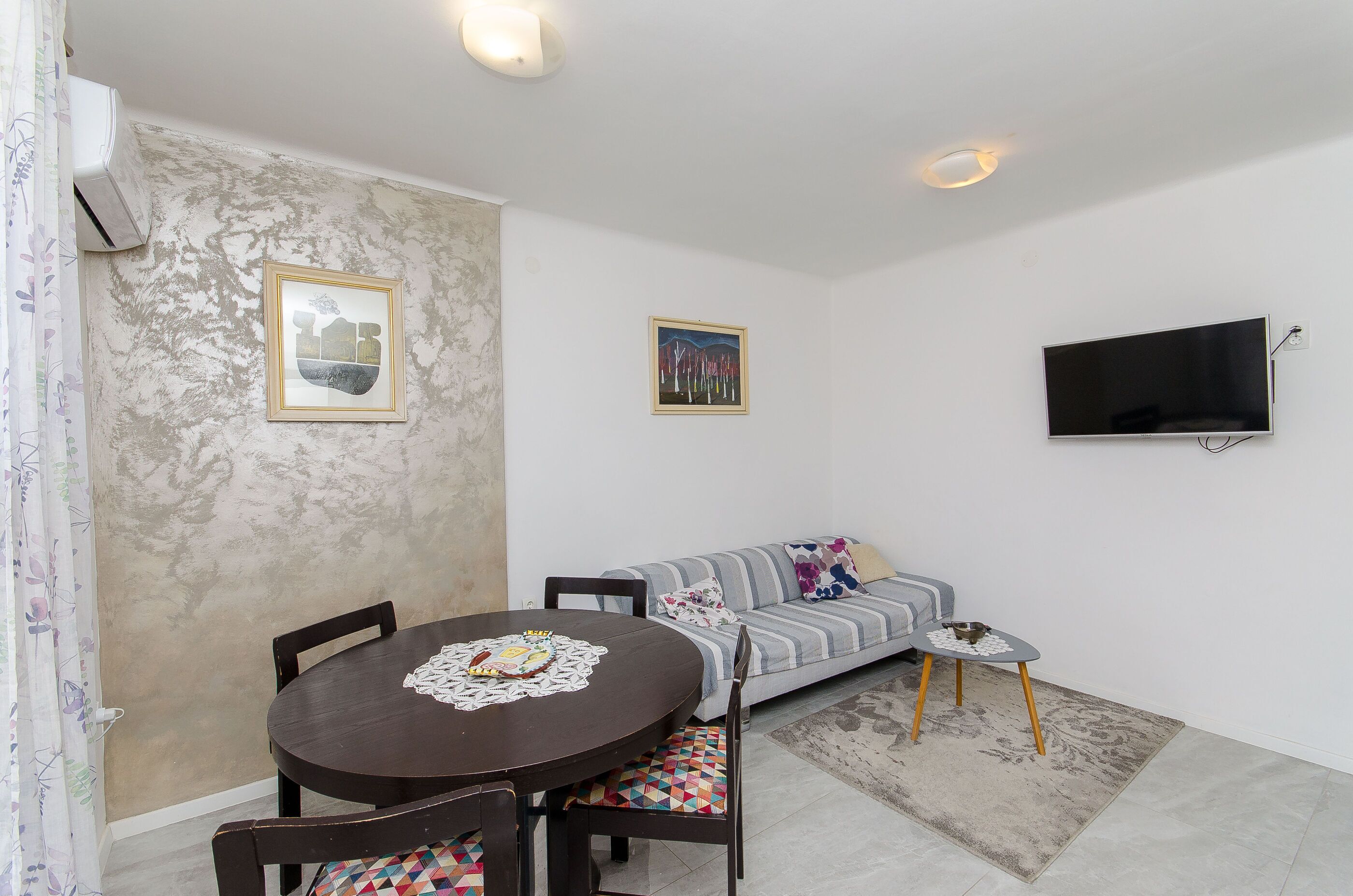 A dining area with a round table and chairs, a sofa, a flat-screen TV mounted on the wall, and a patterned rug on the floor.