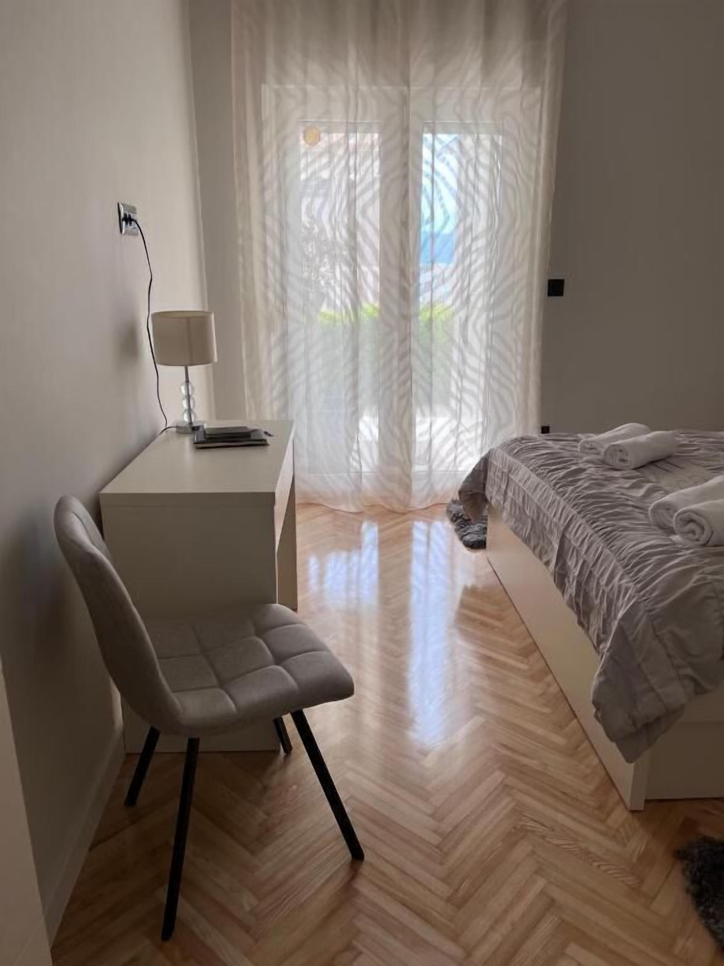 Ein modernes Schlafzimmer mit Holzboden, einem Bett mit gemustertem Überwurf, einem Schreibtisch mit Lampe und durchscheinenden Vorhängen, die natürliches Licht hereinlassen.