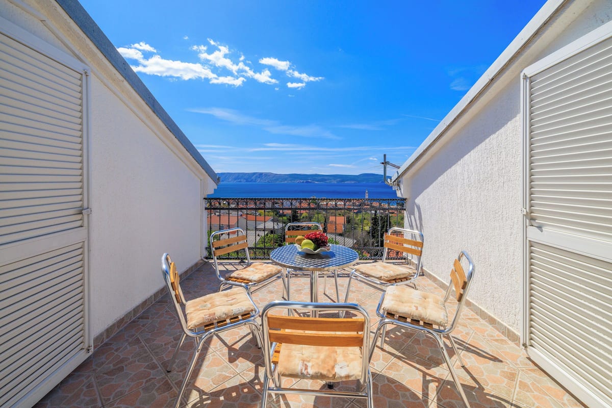 Eine sonnendurchflutete Terrassenanlage im Freien mit einem runden Tisch und sechs Stühlen, Blick auf das Meer und einem Metallzaun.
