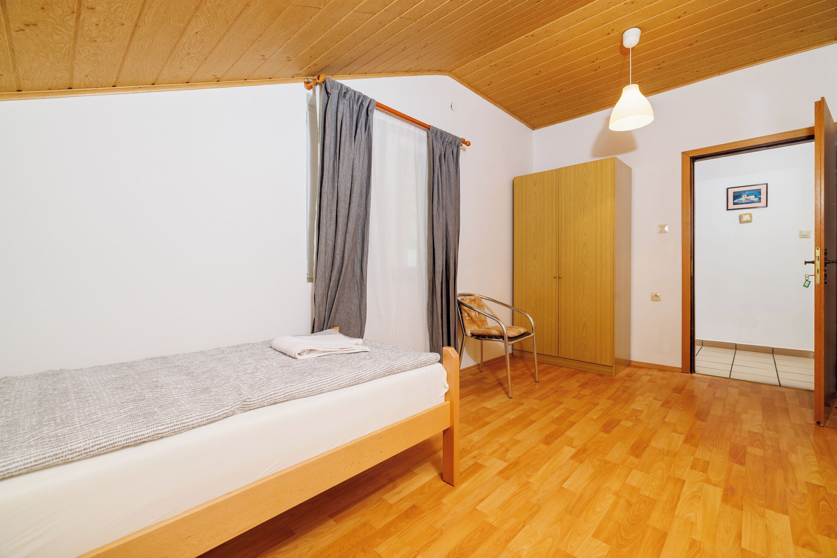 A single bed with a wooden frame, a chair, and a wardrobe in a room with wooden flooring and a sloped ceiling.