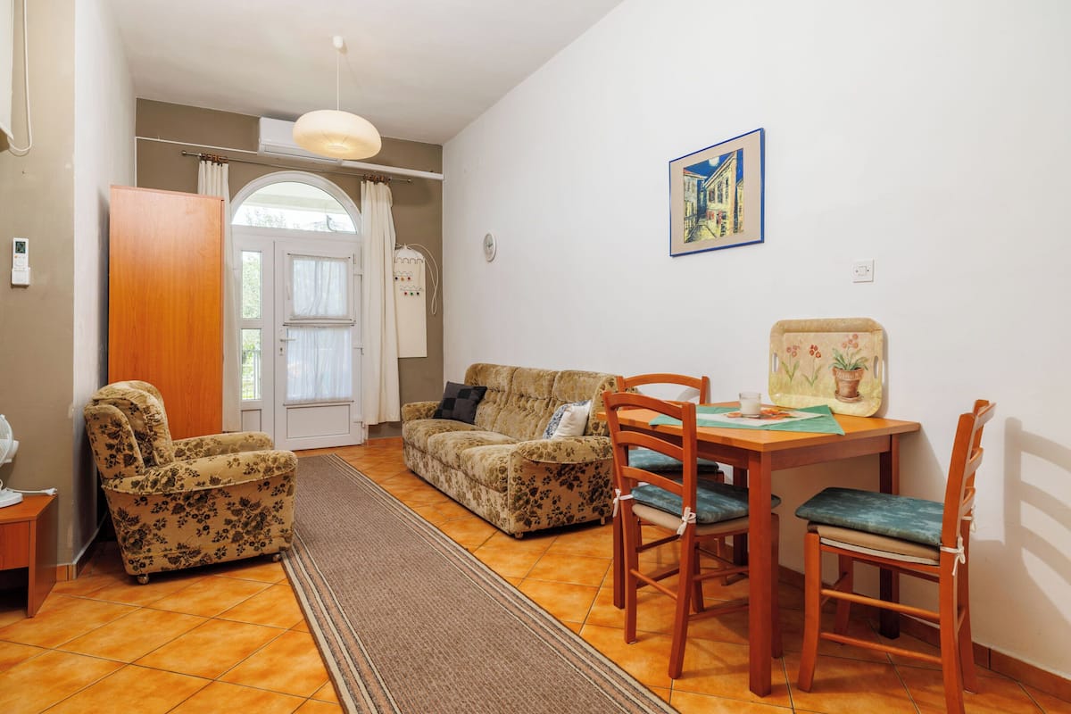 Ein Wohnzimmer mit einer Couch, zwei Sesseln, einem Esstisch mit Stühlen, einem Teppich und einem Bild an der Wand.