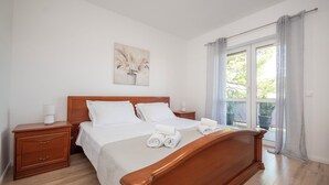 A neatly made bed with white linens and rolled towels, a wooden headboard, a bedside table with a lamp, and a window with curtains. - House Nevenka (Omis)