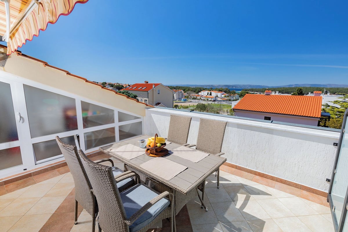 A balcony with wicker furniture, a table with fruits, and a view of houses and mountains.