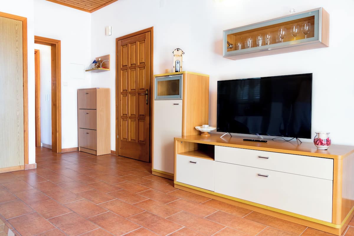 A living room with a TV, wooden cabinets, a glass cabinet with wine glasses, and a tiled floor.