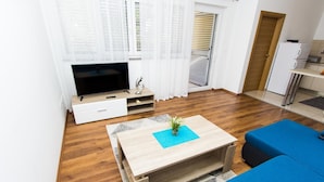 Un salon moderne avec une table basse en bois, un canapé bleu, un téléviseur à écran plat et un coin cuisine équipé d’un réfrigérateur et d’un four à micro-ondes.