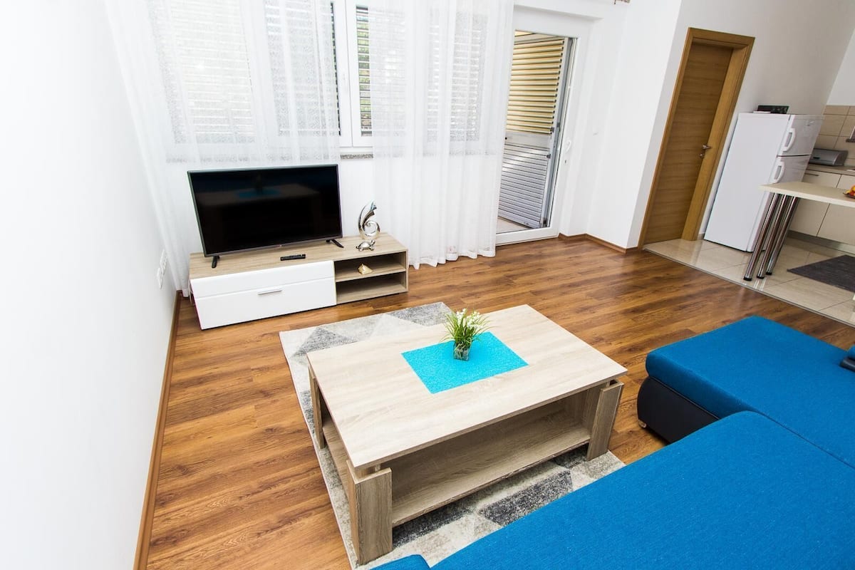 Ein modernes Wohnzimmer mit Couchtisch aus Holz, einem blauen Sofa, einem Flachbildfernseher und einer Küchenzeile mit Kühlschrank und Mikrowelle.