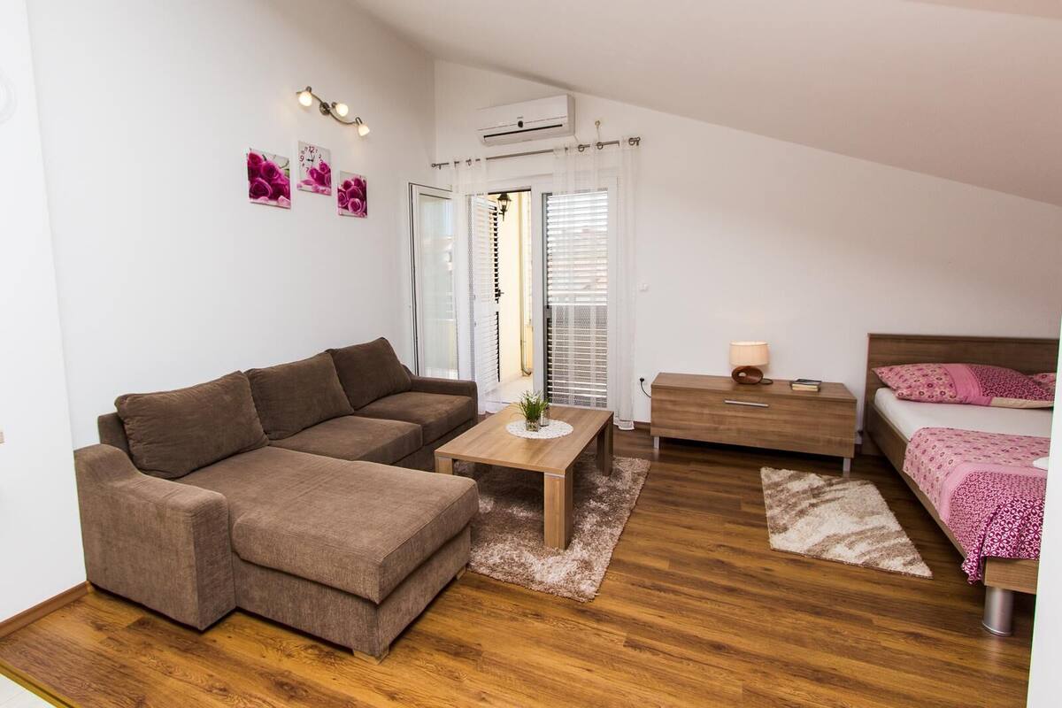 Ein kompakter, moderner Schlafzimmerraum mit einem braunen Sofa, einem Holznachttisch, einem Beistelltisch mit Lampe und einem Bett mit rosa Bettwäsche.