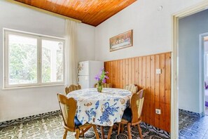 A dining area with a round table, chairs, a vase with flowers, a refrigerator, and a window with curtains. - Apartments Katin Kutak (Sibenik)