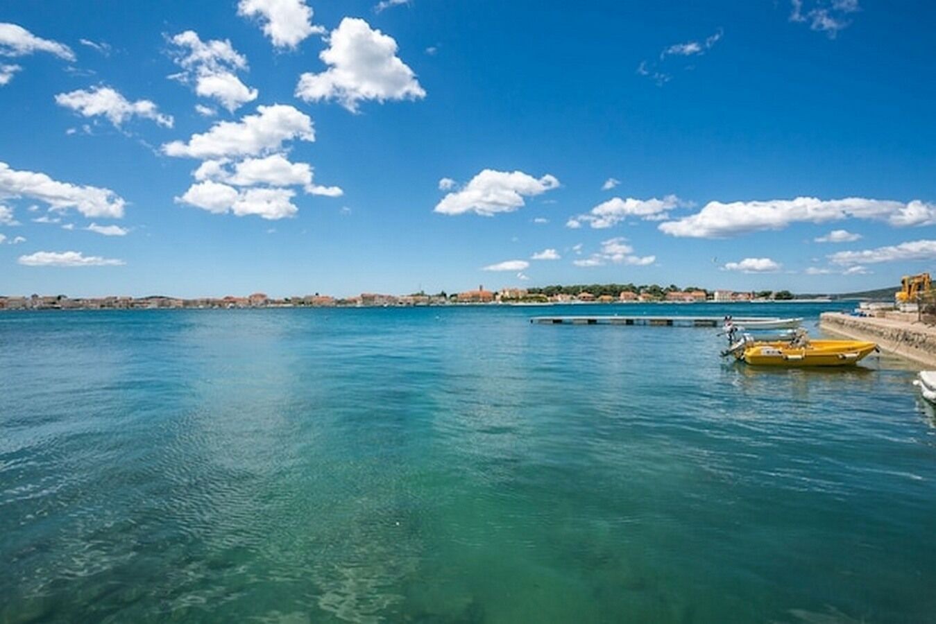 Kustlandschap met helder turquoise water, een gele boot aan de rechterkant en een verre kustlijn met gebouwen.