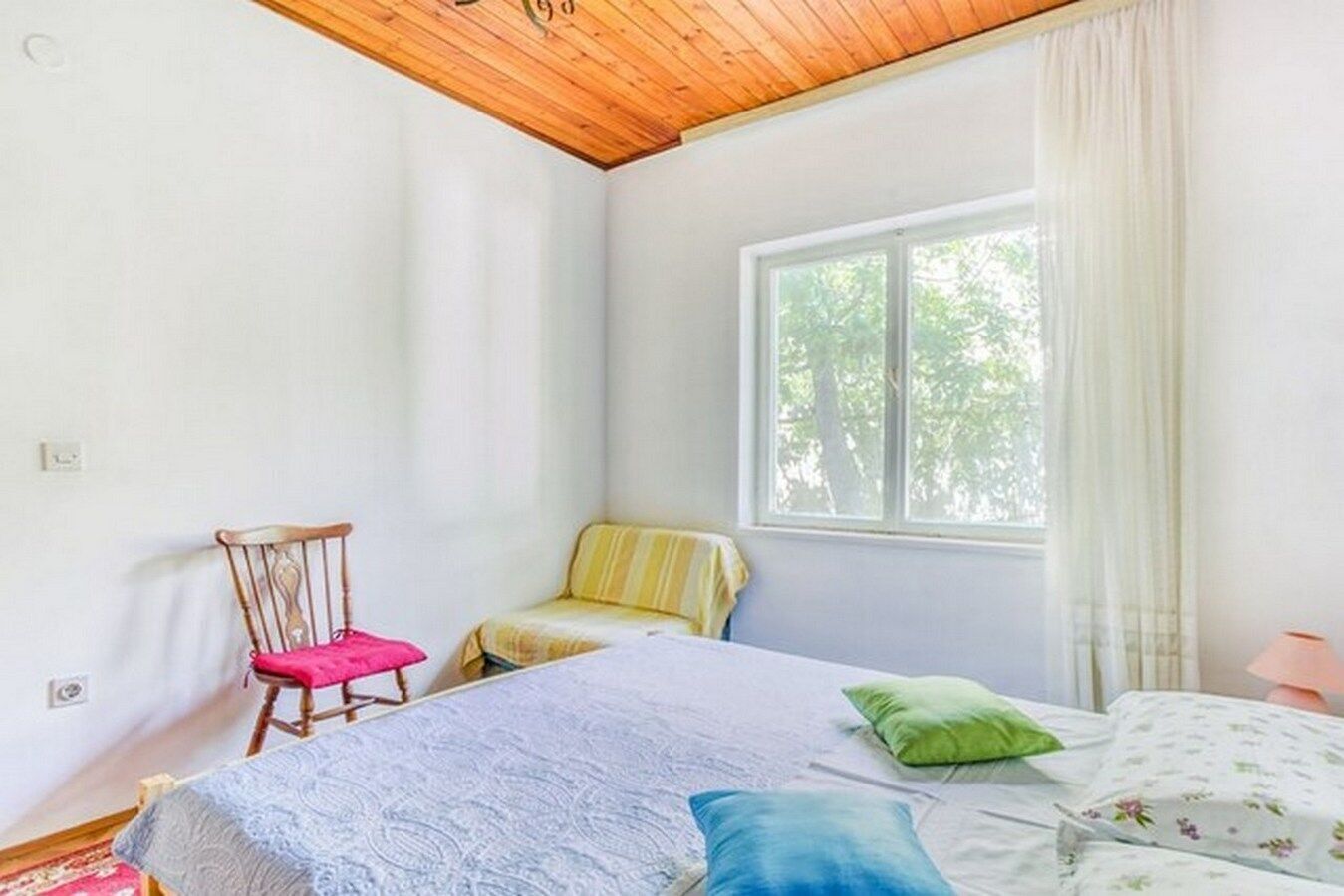 A bedroom with a wooden ceiling, a bed with a white and floral bedspread, a chair with a pink cushion, and a window with white curtains.